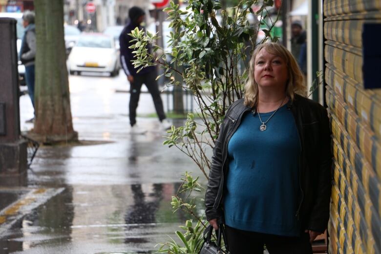 A woman stands in front of a plant, wearing black jacket and blue shirt. 