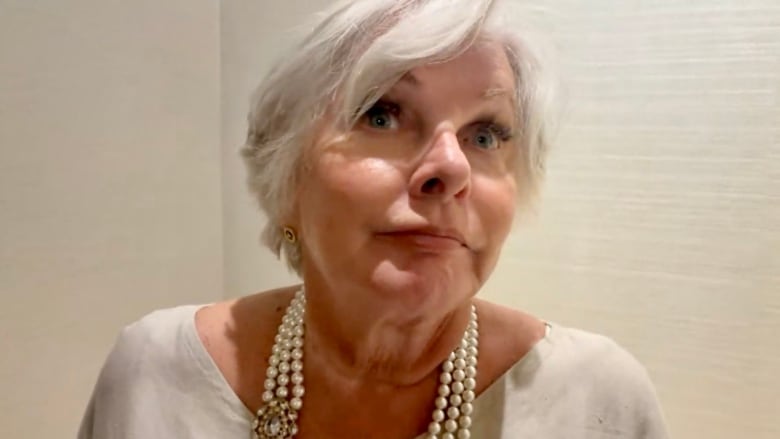 A white-haired woman wearing earrings and a triple-strand pearl necklace is seen in front of an off-white wall.