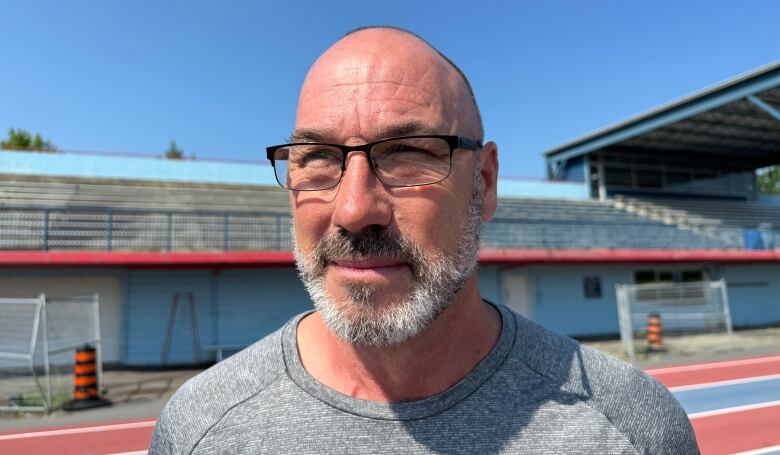 A man with a beard and glasses squints into the sunlight standing in front of a stadium