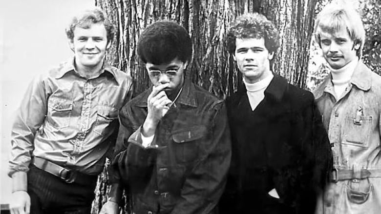 Four men with 70s-era haircuts lean against a tree in a black-and-white photo.