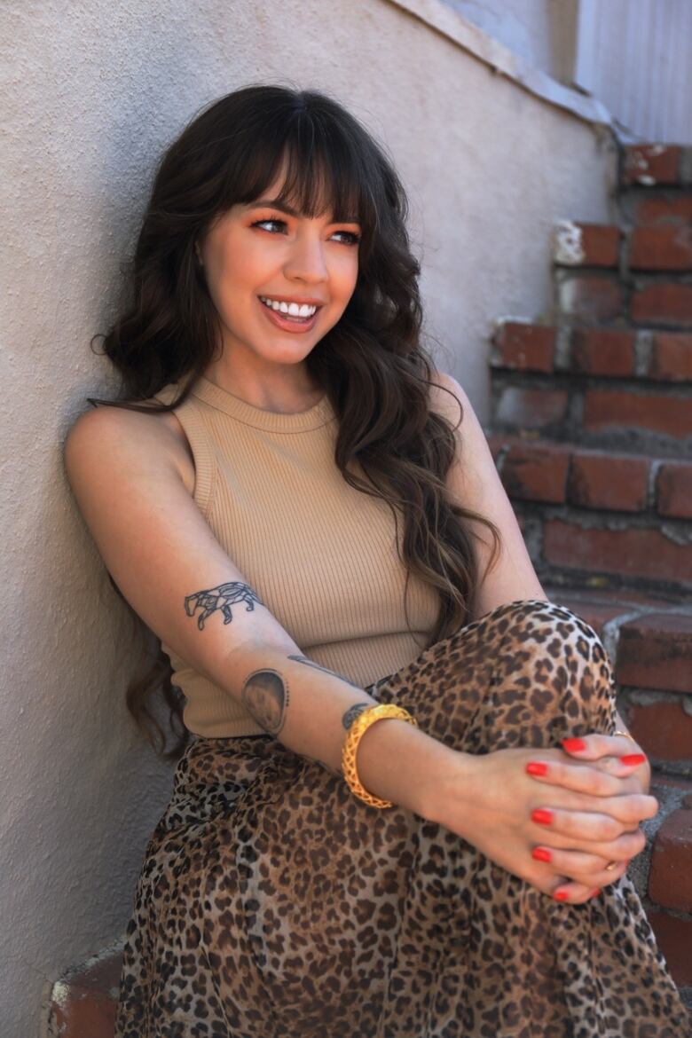 A smiling woman with long brown hair sits on an outdoor brick stairwell with her hands wrapped around one knee.