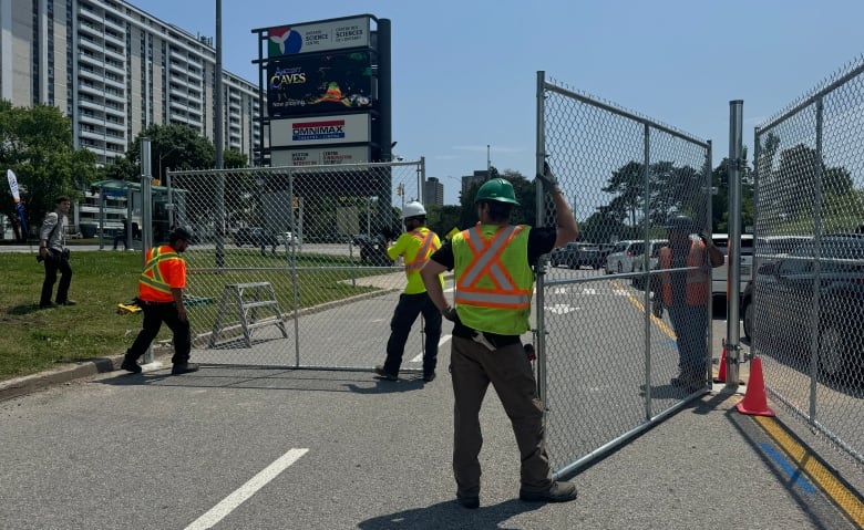 Workers erect a chain-link fence on a road, with an Ontario Science Centre sign in the background.  