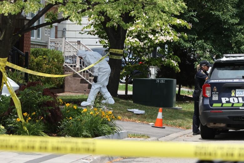 A person in an all white body suit walks underneath crime scene tape set up in the front yard of a suburban home.