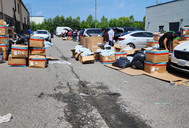 Dozens of boxes scattered around a parking lot, blocking the lane for cars. 