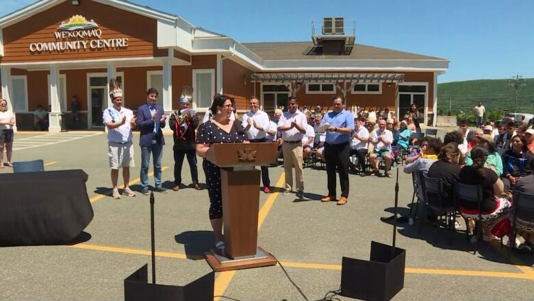 A woman with dark hair wearing a polka dot dress stands at a podium in front of a community centre. People behind her are clapping.