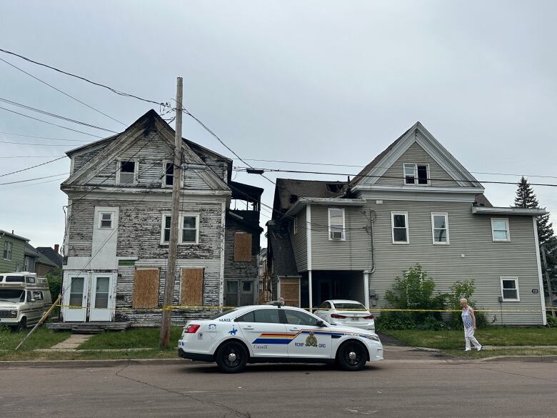 Yellow police tape stretches across the front of two houses, one with boarded up windows. The tops of both buildings are blackened from fire damage. A police cruiser is parked in front.