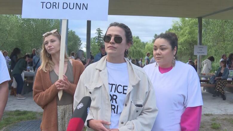 Three women look at a camera at a vigil. The person in the centre is wearing a T-shirt reading 'Justice for Tori', with a woman to the left carrying a sign that partially reads 'Tori Dunn'.