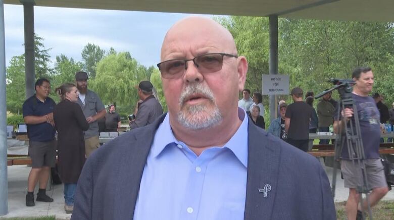 A bald white man speaks at a vigil, wearing a blue shirt and a navy blue coat.