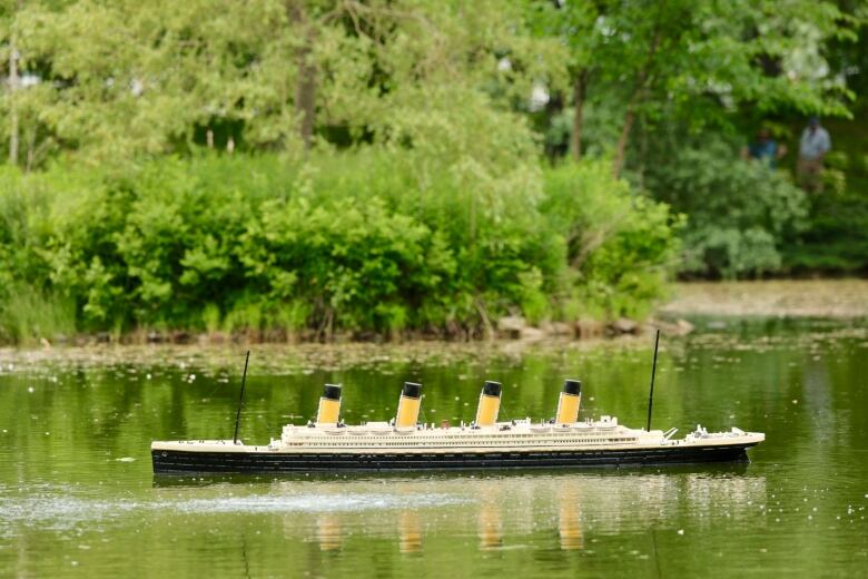 A replica of the Titanic floats in a body of water with the bank of a pnd in the background.