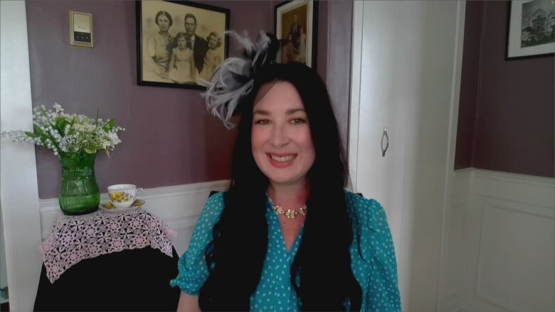 A smiling woman with long black hair wearing a blue dress and a fascinator sits in a room with purple walls. The back wall has a photo of Queen Elizabeth II and her parents on it.