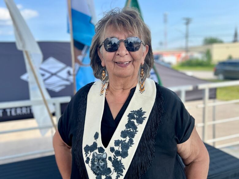 a woman with brown and grey hair wears a black t shirt, sunglasses, and orange and black beaded earrings and smiles for the camera