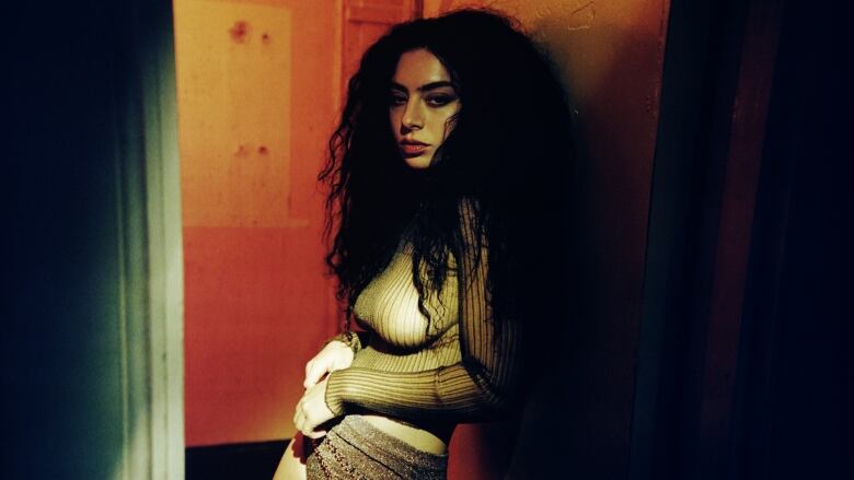 A woman with big dark hair in a striped long sleeve top and short sparkly shorts stands in a dimly lit doorway.