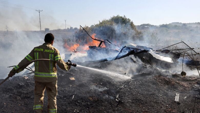 A man wearing firefighting gear uses a hose to spray water on a pile of burning, smoking rubble in a field.  