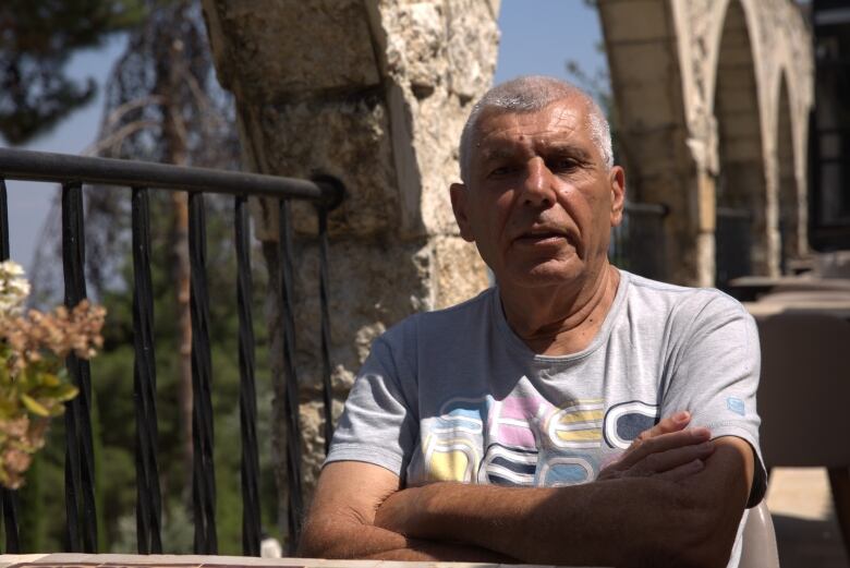 A man with close-cropped grey hair wears a t-shirt and folds his arms across his chest while sitting in a courtyard. 