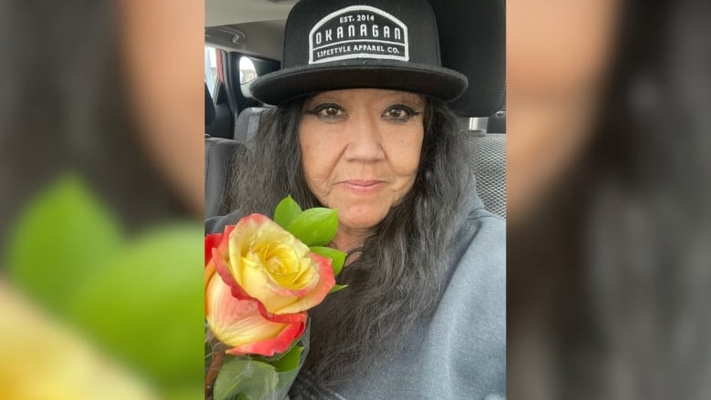 An Indigenous woman poses for a selfie while wearing a black hat and carrying flowers.