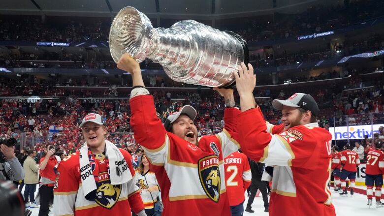 Flanked by two other players, a hockey player in a baseball cap lifts a huge silver trophy over their head. 