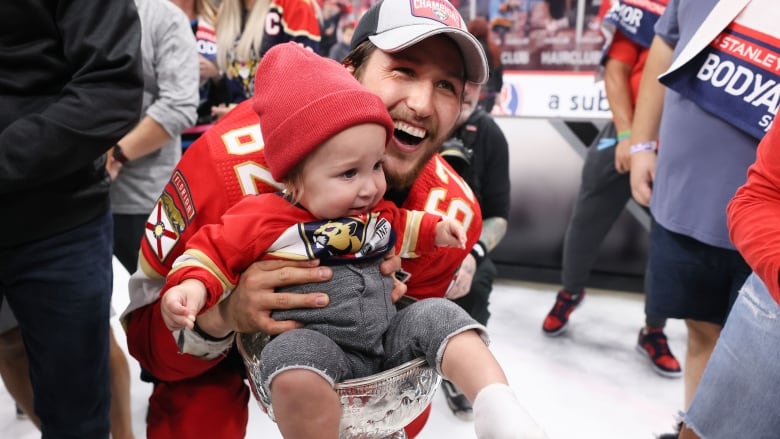 A smiling hockey player in a baseball cap crouches and holds a baby whose bottom is in the top of a silver trophy. 