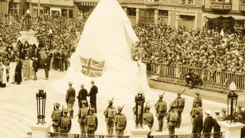 A black and white photo of a large gathering of people surrounding a statue covered by white canvas. 