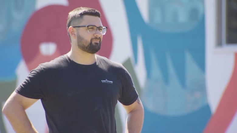 A man with dark hair and glasses looks away from the camera on a sunny day.