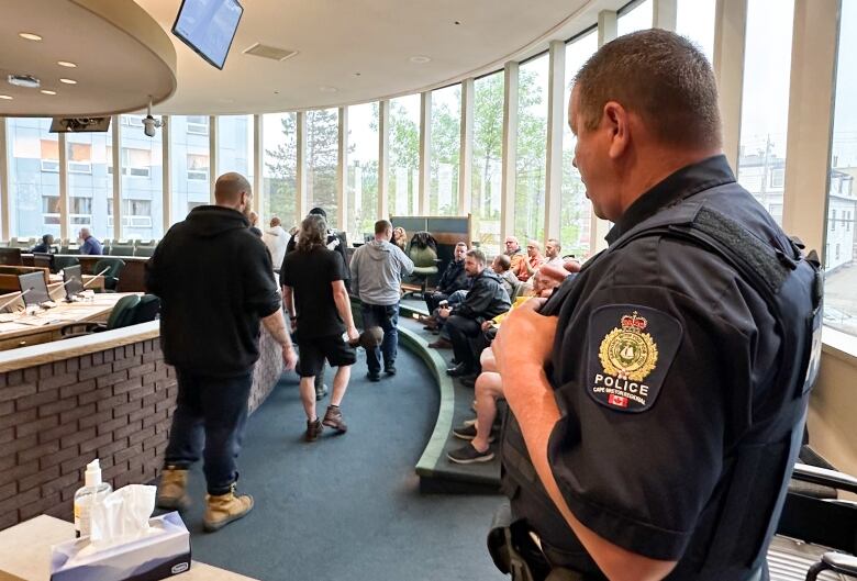 A police officer stands in the foreground with his back to the camera watching as a number of people walk into a room with very tall windows.