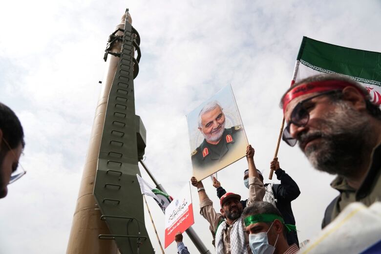 A demonstrator holds aloft a placard bearing the image of a man in a military uniform. Another man behind him waves a large Iranian flag. Both are standing next to a thin tower. 