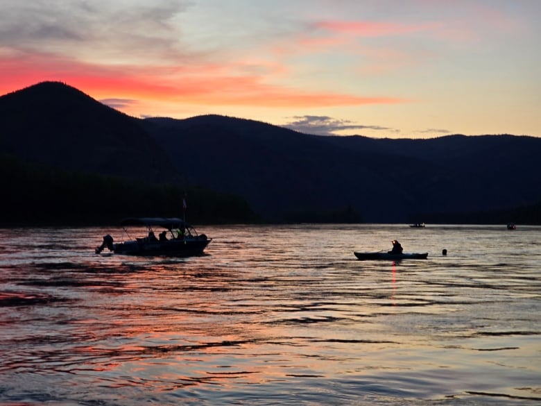 A boat and kayak are seen in silhouette near a swimmer in the water at twilight.