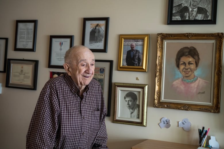 Joseph Novak at the Whistle Bend Place Care Facility in Whitehorse, Yukon. He is an older white man who is smiling alongside a range of pictures, paintings and memorabilia.