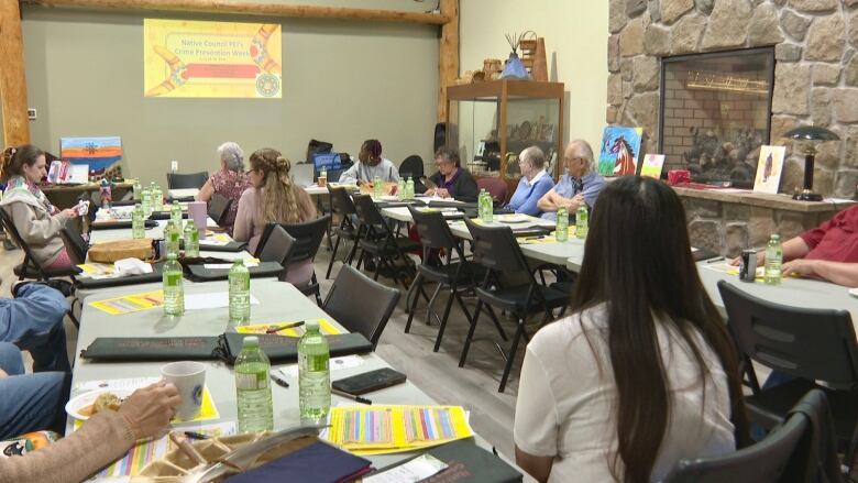 A room with two long grey tables. They are covered in green water bottles and yellow paper. About a dozen people are sitting looking towards the  front of the room. It says Crime Prevention Week on the wall.