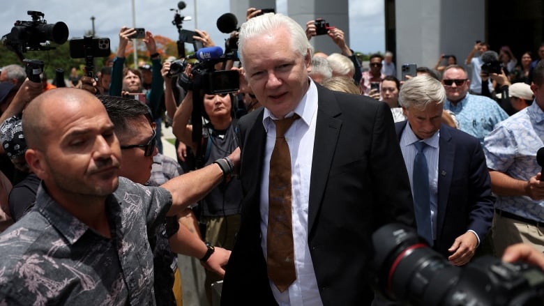 A man with grey hair and wearing a black suit walks amid a throng of people.