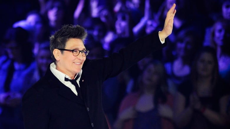 A woman with short black hair and glasses, wearing a shirt, tie and cardigan, waves at the crowd.