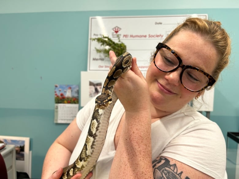 A woman with long hair in a ponytail and thick, dark-framed glasses holding a small ball python.