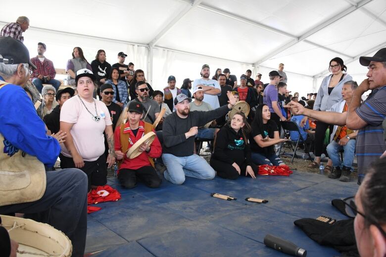 People kneel on a blue mat in a white tent. Some of them are holding drums. 