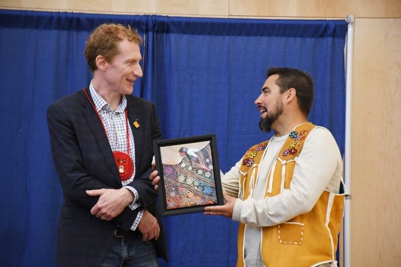 A man in a dark suit looks at a man wearing a beaded vest holding a picture. 