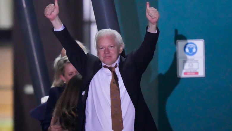 A man with slicked-back white hair, wearing a dark suit jacket, white collared shirt and brown silk tie, gives two thumbs up high above his head as he walks out of a building.