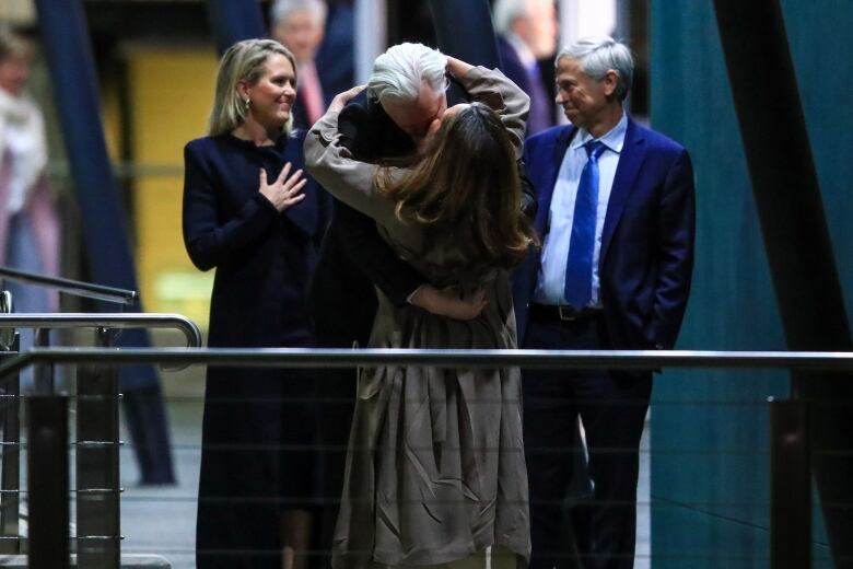 A man with white hear embraces and kisses a woman as another woman, on the left, and a man, on the right, smile in the background. 