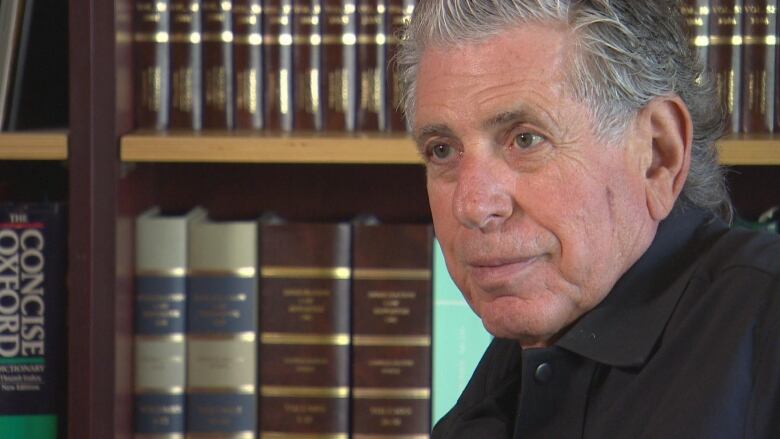 A man in a black collared shirt is seated in front of a book shelf. 