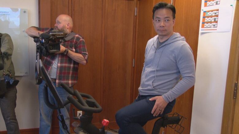 A man rides a stationary bicycle as journalists watch.