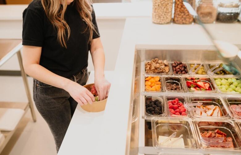 A woman assembles a bowl while toppings of fresh vegetables, fruit and meat can be seen in clear plastic containers next to her.