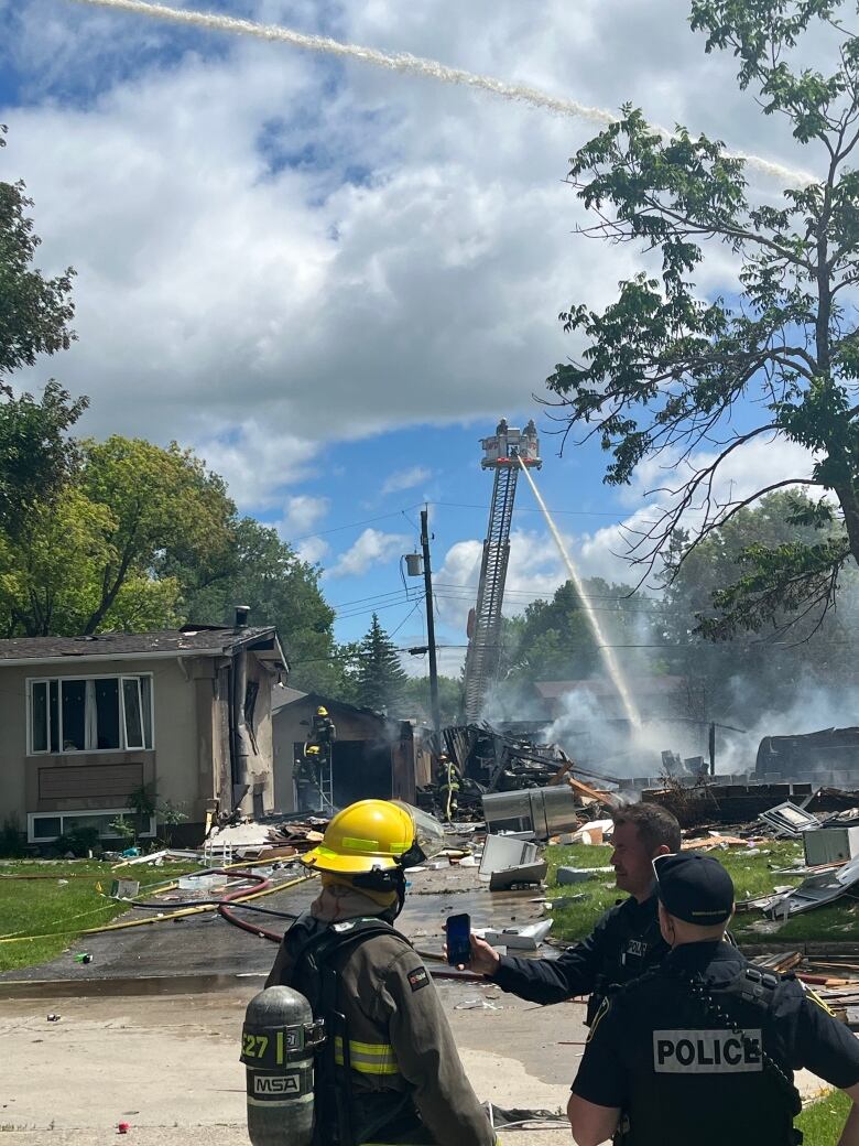 A damaged house next to a pile of rubble. Firefighters work nearby.