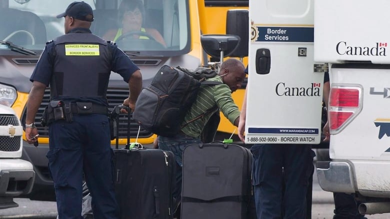 An officer and another man unload suitcases from a vehicle.