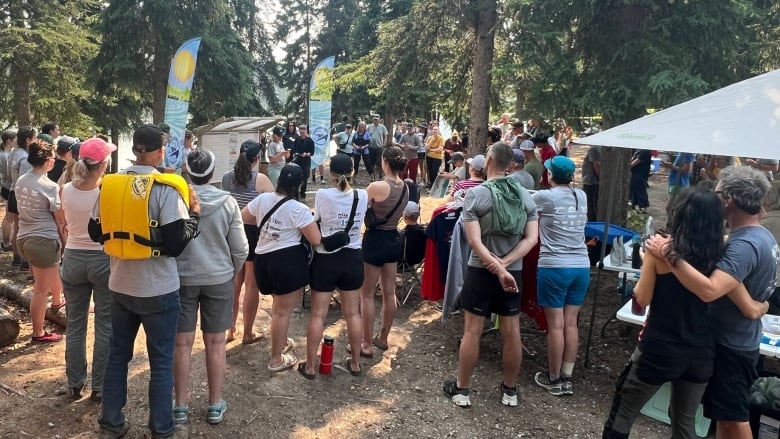 A group of people stand in a wooded area listening to someone talk.