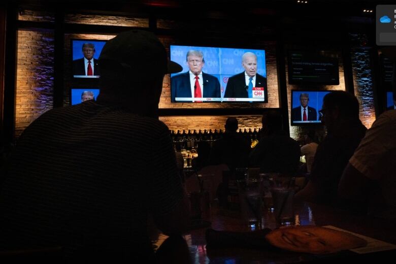 People at a bar watch a debate on multiple wall-mounted televisions.