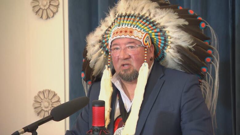 A man in a headdress speaks at a podium 