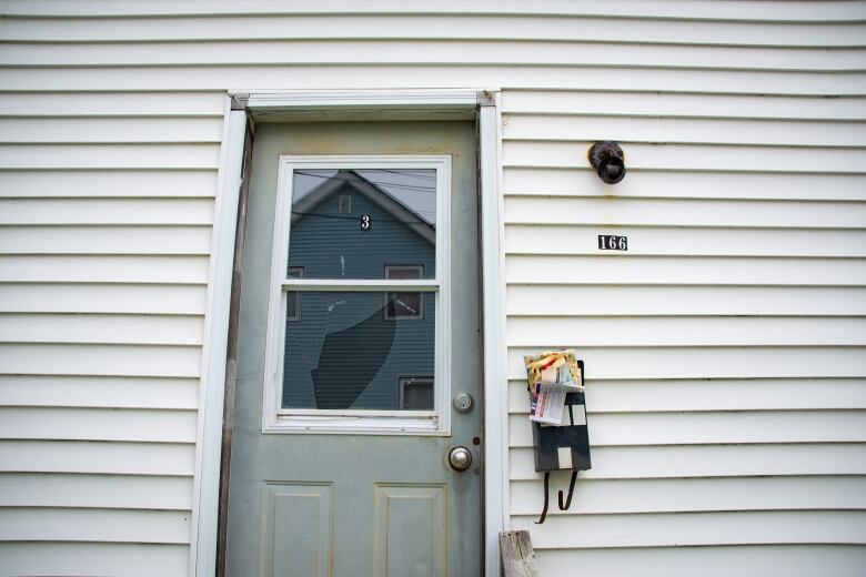 A window is smashed in a metal door that is covered in mildew, next to an overflowing black mailbox.