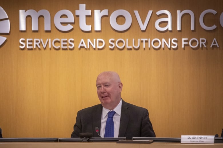 A man in a suit sits at a desk with a Metro Vancouver sign behind him.