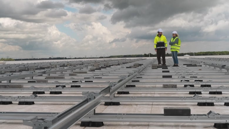 Susan Ormiston stands on a roof with a Sunrock project manager, looking at a solar panel installation in progress