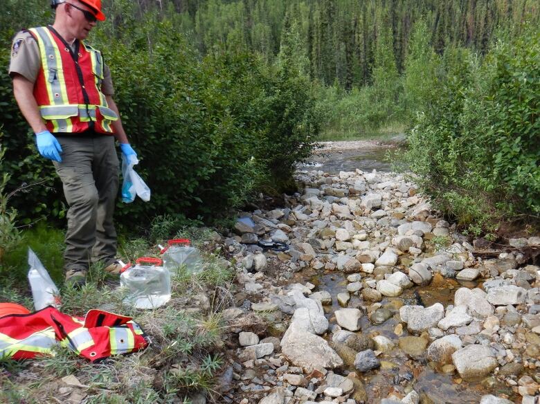 A man in a safety vest stands by a small creek with some water jugs.