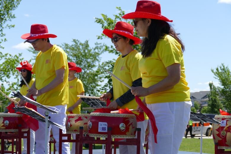 People wearing yellow shirts and red hats drum at an event. 