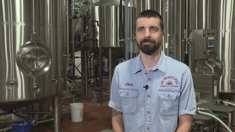 A man in a denim shirt stands in a room full of silver brewing equipment.