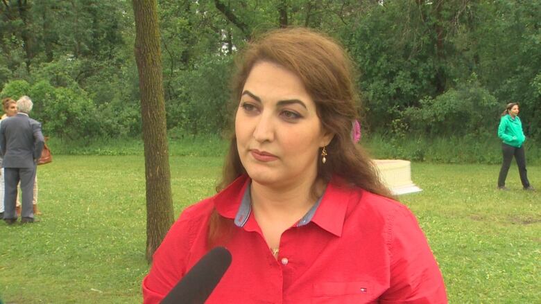 A woman with a red shirt stands in front of a forest backdrop.